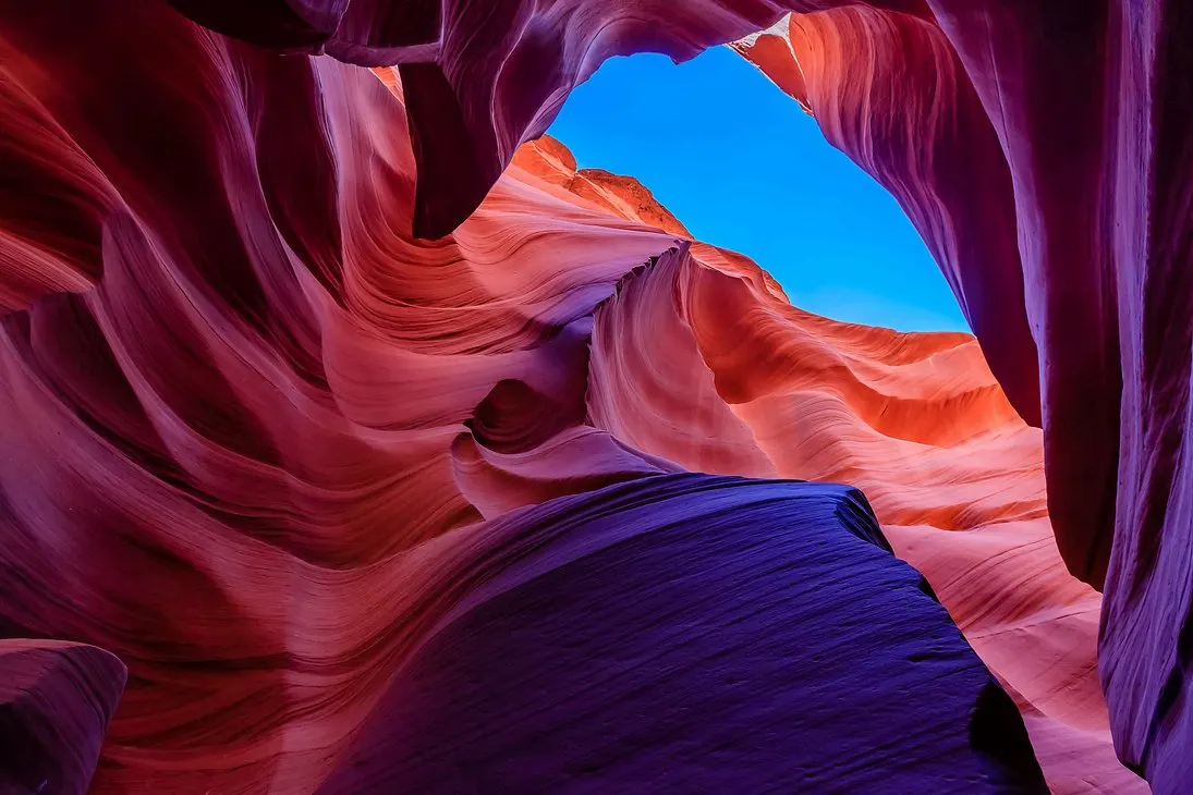 El interior de Antelope Canyon, con las paredes de arenisca ondulada en tonos naranjas y morados y una franja de cielo azul en lo alto.
