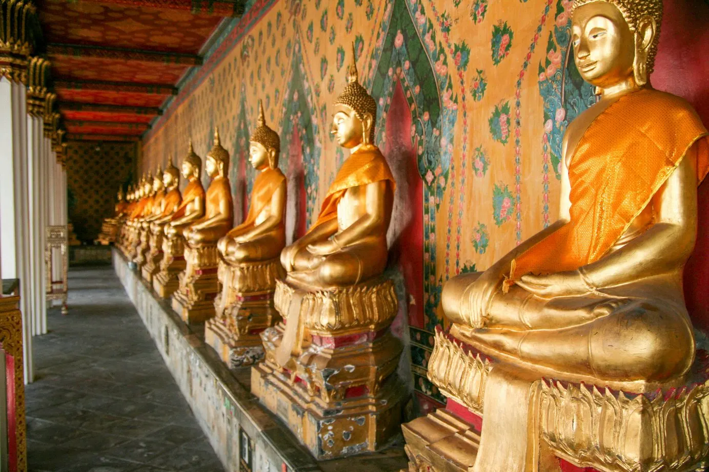 Hilera de budas dorados sentados en la galería de un templo de Bangkok, sobre pedestales y frente a un muro de mosaicos de colores.