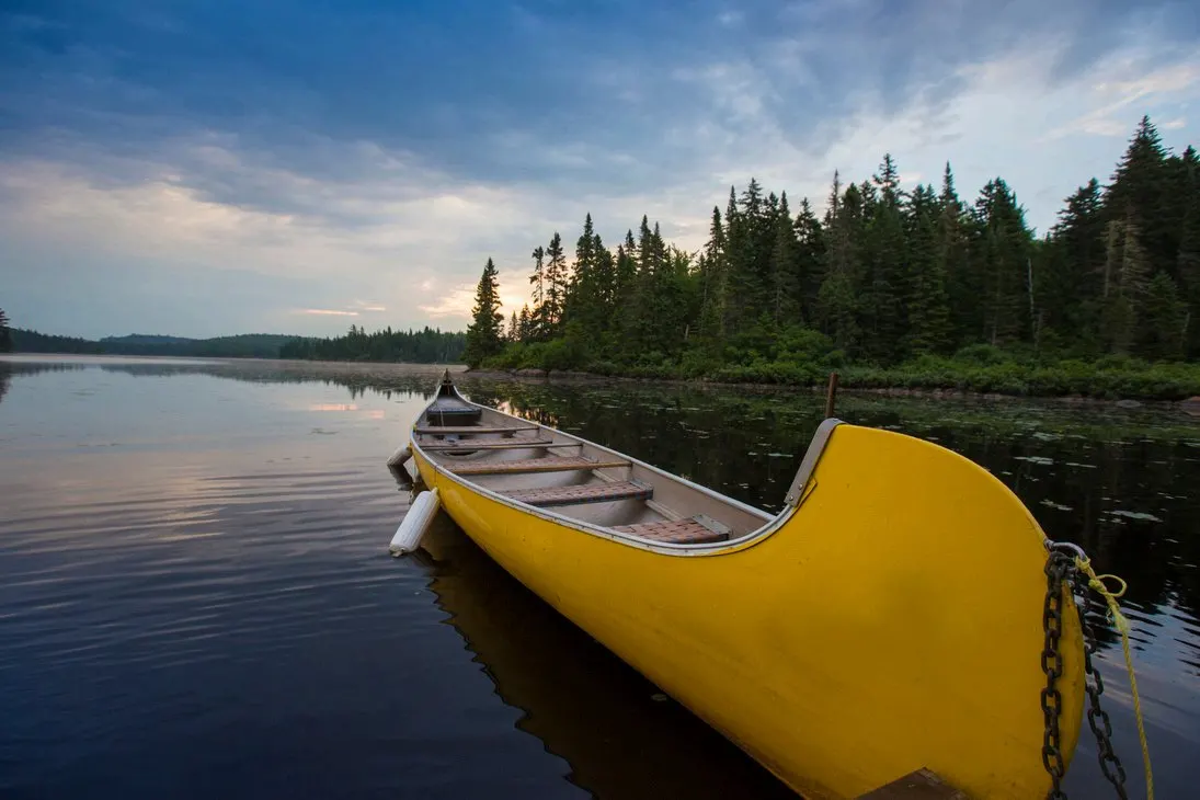 Una canoa amarilla amarrada en la orilla de un lago en calma, con el bosque de coníferas y el cielo del atardecer al fondo.