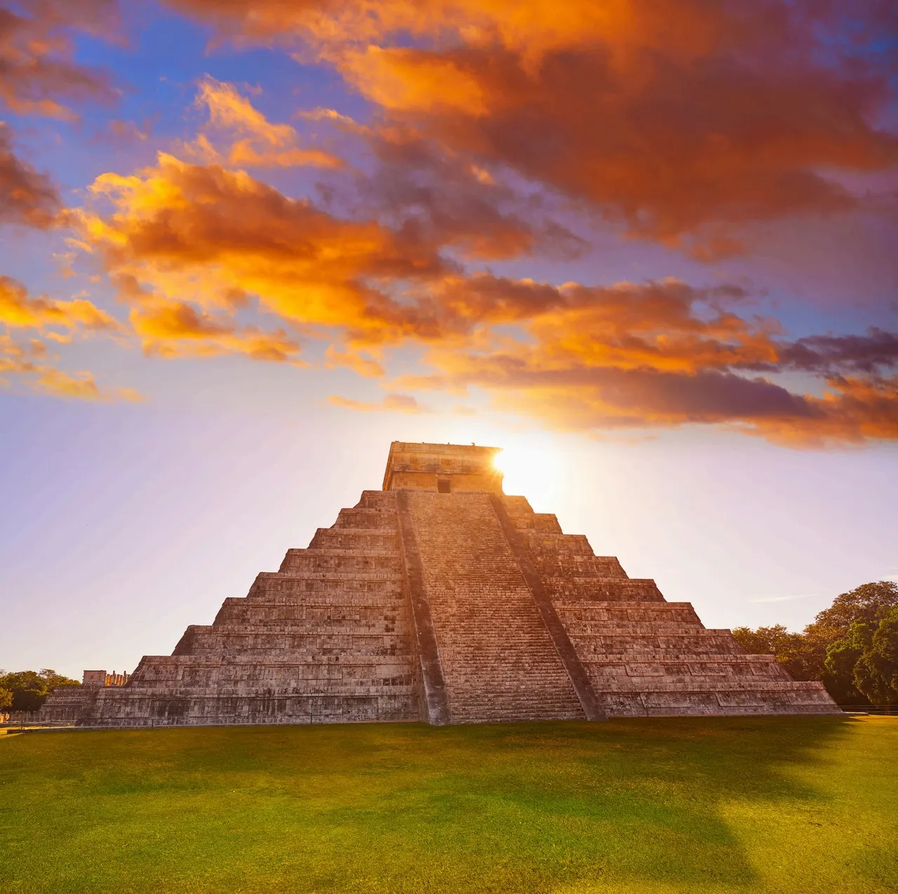 La pirámide de Kukulkán de Chichén Itzá al atardecer, con el sol asomando tras la escalinata y nubes anaranjadas sobre la explanada de hierba.
