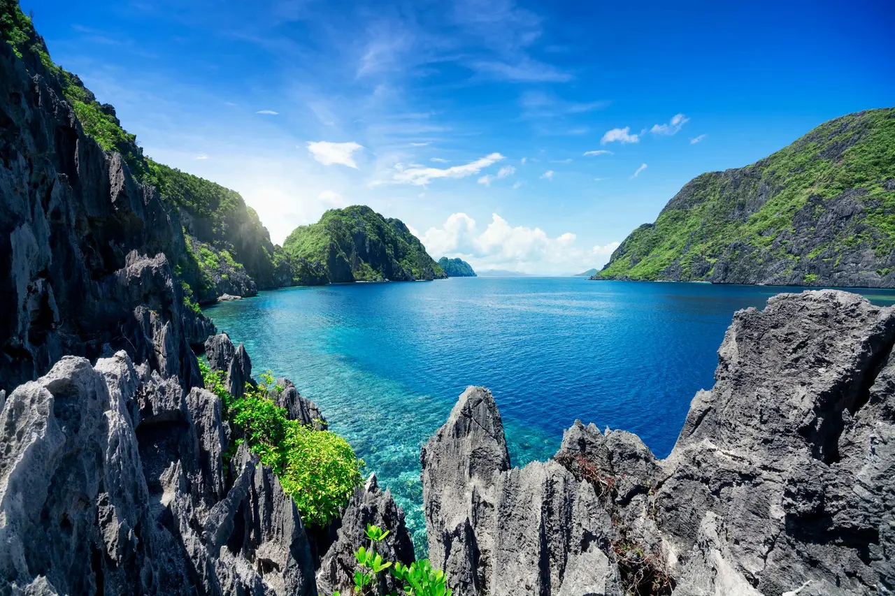 Una bahía de El Nido vista entre rocas kársticas afiladas en primer plano, con el agua turquesa y las islas cubiertas de vegetación al fondo.