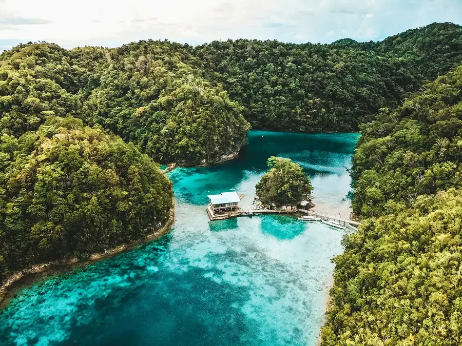 Vista aérea de una laguna de agua turquesa encajada entre colinas cubiertas de selva, con una plataforma de madera sobre el agua en el centro.