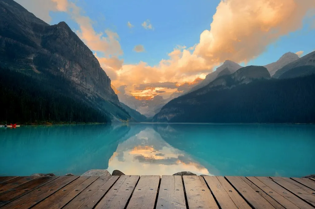 El lago Louise al amanecer desde un embarcadero de madera, con las montañas y las nubes anaranjadas reflejadas en el agua turquesa.