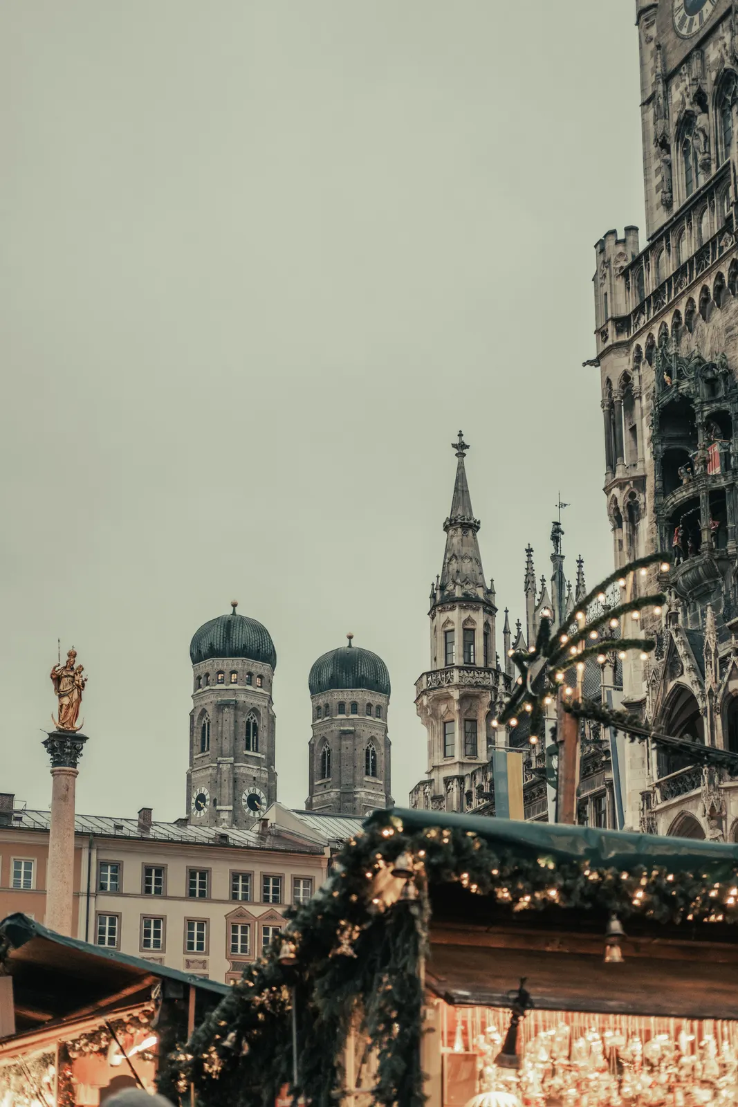 Casetas de madera del mercado navideño de la Marienplatz de Múnich, Alemania, con guirnaldas de abeto, luces y campanillas; al fondo, la columna de la Virgen, las dos torres de cúpula de bulbo de la Frauenkirche y la torre del Nuevo Ayuntamiento