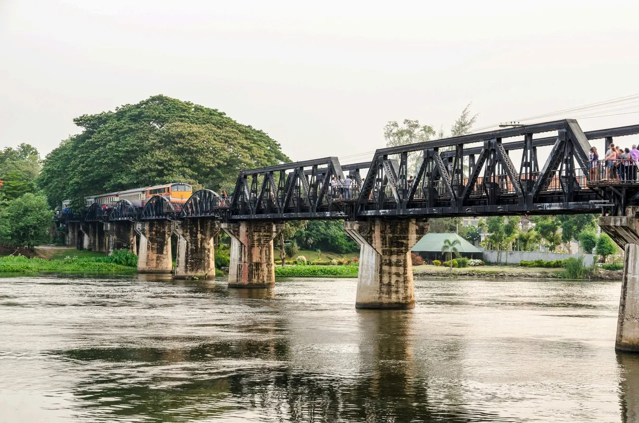 El puente ferroviario sobre el río Kwai, con un tren cruzando su estructura metálica negra y el río discurriendo entre los pilares de hormigón.