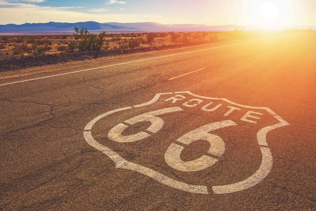 El escudo de la Ruta 66 pintado en blanco sobre el asfalto de una carretera recta del desierto, con el sol bajo en el horizonte.