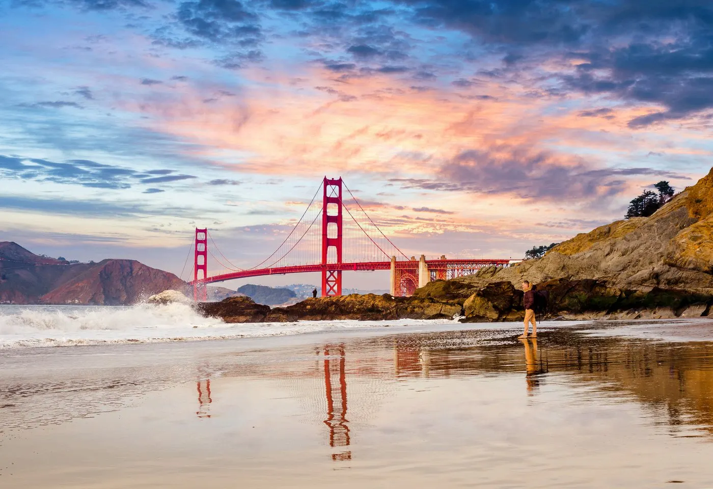 El puente Golden Gate de San Francisco visto desde la playa al atardecer, con el cielo rosado reflejado en la arena mojada.
