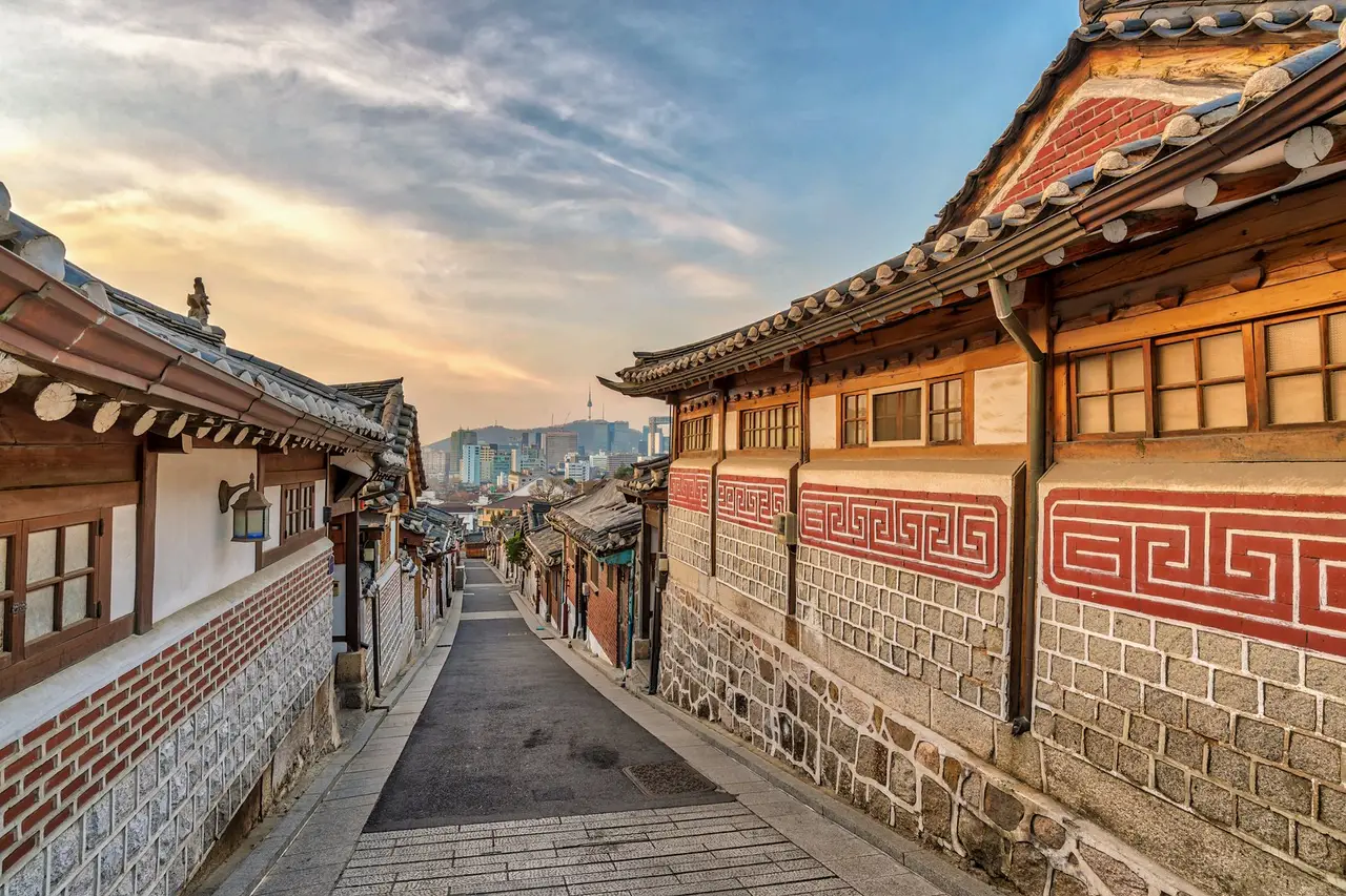 Una calle estrecha de casas tradicionales hanok en Seúl, con los tejados curvos de teja y las celosías de madera, y la torre de la ciudad al fondo.
